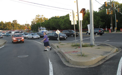 Photo shows a right-turning lane at a corner where one street intersects a 6-lane street that has traffic waiting for a green light.  The right-turning lane is separated from the rest of the street by an island, and a woman has just left that island to cross the right-turning lane to reach the sidewalk on the corner.  There is a car in the lane waiting for her.